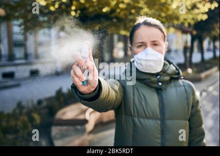 Ritratto di una donna all'aperto con maschera monouso e disinfettante spray per disinfettare le mani alla fotocamera. Bella giornata di primavera durante la quarantena di coronavirus. Concetto di distanza sociale Foto Stock