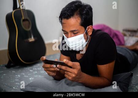 Un giovane uomo con maschera chirurgica stava giocando a giochi mobili a casa sua a causa della quarantena domestica del coronavirus a Dhaka. Foto Stock