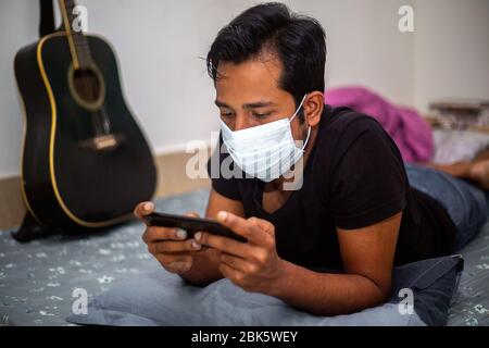Un giovane uomo con maschera chirurgica stava giocando a giochi mobili a casa sua a causa della quarantena domestica del coronavirus a Dhaka. Foto Stock