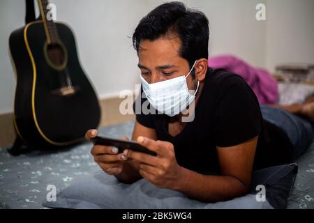 Un giovane uomo con maschera chirurgica stava giocando a giochi mobili a casa sua a causa della quarantena domestica del coronavirus a Dhaka. Foto Stock