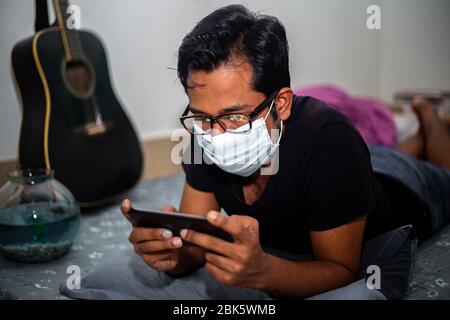 Un giovane uomo con maschera chirurgica stava giocando a giochi mobili a casa sua a causa della quarantena domestica del coronavirus a Dhaka. Foto Stock