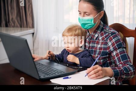 Rimani a casa madre in una maschera che lavora da remoto mentre si prende cura del suo bambino. Le tecnologie moderne contribuiscono a mantenere il lavoro. Messa a fuoco selettiva Foto Stock