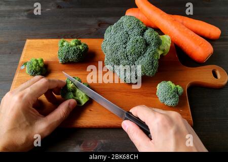 Mano che taglia broccoli crudi su legno tagliando asse con le carote sfocate nello sfondo Foto Stock