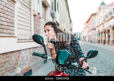 Giovane bella donna mettere rossetto guardare in specchio, mentre seduto sul suo scooter. Foto Stock