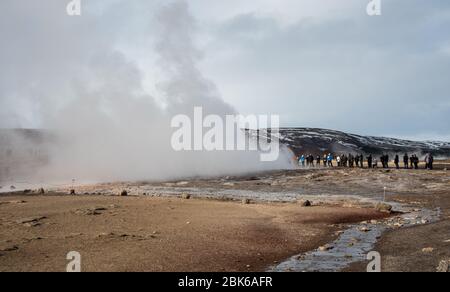 Persone che guardano all'esplosione di acqua bollente calda ai geyser nella zona geotermica di haukadalur in Islanda Foto Stock