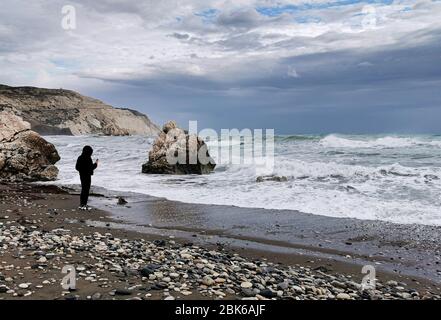 Persona sconosciuta che gode il mare tempestoso con le onde ventose. Luogo di nascita della roccia di Afrodite, costa di Petra tou romiou nella zona di Paphos a Cipro Foto Stock