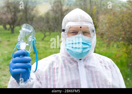 Ritratto di uomo anziano che indossa abiti, occhiali, maschera e guanti di nocciola sta tenendo una maschera di ossigeno all'aperto Foto Stock