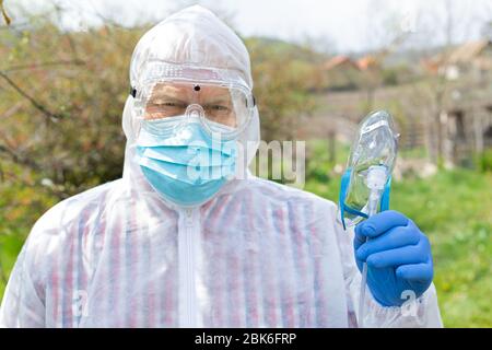 Ritratto di uomo anziano che indossa abiti, occhiali, maschera e guanti di nocciola sta tenendo una maschera di ossigeno all'aperto Foto Stock