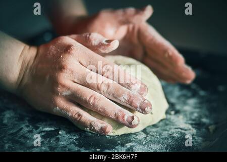 Il cuoco sta impastando la pasta di pane con mani forti su una teglia scura ricoperta di farina bianca friabile, illuminata dalla luce. Foto Stock