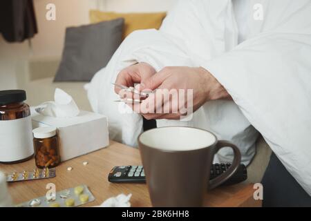 Mani di giovane uomo malato sotto coperta seduto da un piccolo tavolo con varietà di medicina e andando prendere la compressa whle rimanere a casa Foto Stock