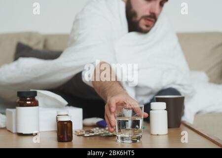 Giovane uomo malato avvolto in coperta seduta sul divano e prendendo il vetro di acqua dal tavolo mentre andare a prendere painkiller o altro medicinale Foto Stock