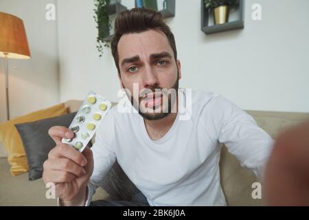Uomo al bearded con influenza, freddo o covid19 che mostra blister con compresse al medico durante la consultazione online di fronte al gadget mobile a casa Foto Stock