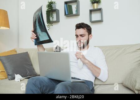 Giovane uomo bearded con fazzoletto e polmoni x-ray risultato seduto sul divano di fronte al laptop e di consulenza con il medico online a casa Foto Stock