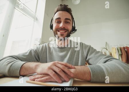 Buon giovane operatore barbuto con un sorriso soothy che ti guarda durante la consultazione online di fronte al notebook in ambiente domestico Foto Stock