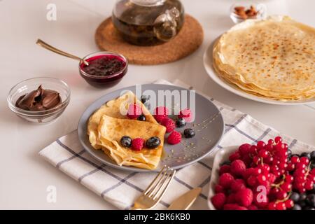 Due pancake piegati con miele e frutti di bosco sul piatto circondati da piccole ciotole di vetro con cioccolato spalmato e marmellata sul tavolo servito Foto Stock