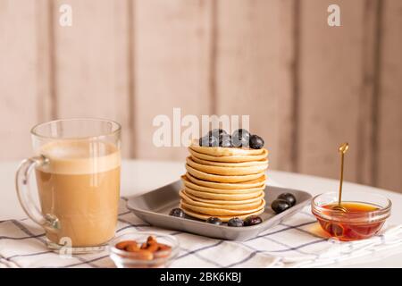 Una pila di appetitosi pancake fatti in casa con more sulla sua cima circondata da un bicchiere di latte e due ciotole con miele e mandorla Foto Stock