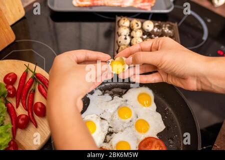 Mani di femmina che rompono le uova fresche di quaglia in padella calda mentre cucinano la colazione con le verdure nella cucina Foto Stock