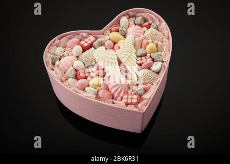 Scatola a forma di cuore rosa riempita con caramelle e marshmallow diversi su uno sfondo nero. Foto Stock