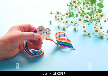 Giornata internazionale infermieri grazie card. Nastro arcobaleno e cuore di legno in mano femminile. Testo "giornata internazionale dell'infermiera" sul cuore. Camomilla fresca Foto Stock
