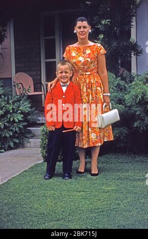 Un ragazzo e sua madre posano sul prato anteriore, indossando abiti eleganti e colorati, USA 1959. Il suo abbigliamento include una giacca rossa brillante e una cravatta ad arco – porta una matita. La donna indossa un abito arancione floreale brillante. Foto Stock