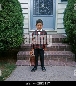 Un ragazzo indossa abiti intelligenti e porta una borsa da scuola, USA c. 1960. Il suo abbigliamento include una giacca da assegno/tartan e una cravatta ad arco. Foto Stock