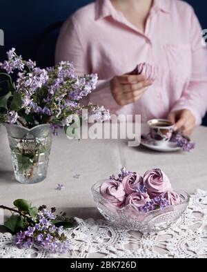 Donna che beve caffè con viola dolce fatto in casa Zephyr o Marshmallow da ribes nero vicino ai fiori lilla su tela grigia da tavolo e sfondo scuro Foto Stock