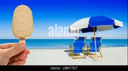 mano con una fresca pop-cicla di alghe davanti a lettini e un ombrellone sulla spiaggia di sabbia argentata Foto Stock