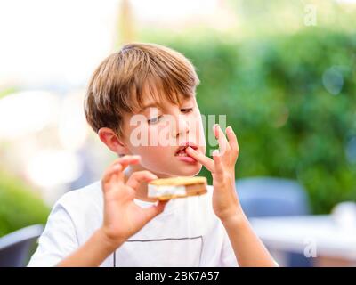 Primo piano ritratto di bel ragazzo leccando dito mentre tenendo e mangiare panino gelato dolce e guardando il gelato Foto Stock