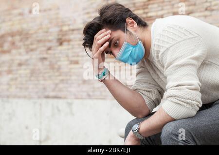 Covid19, giovane preoccupato seduto e fissando al suolo. All'aperto con mano sulla testa con atteggiamento triste. Dispositivi di protezione individuale per coronavirus Foto Stock