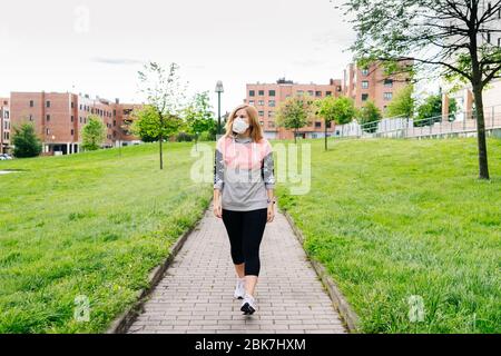 Una donna con una maschera sul viso cammina attraverso un parco con vegetazione verde in un'area residenziale con edifici sullo sfondo Foto Stock