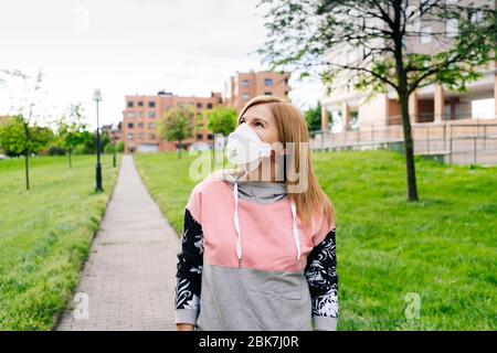 Una donna con una maschera sul viso cammina attraverso un parco con vegetazione verde in un'area residenziale con edifici sullo sfondo Foto Stock