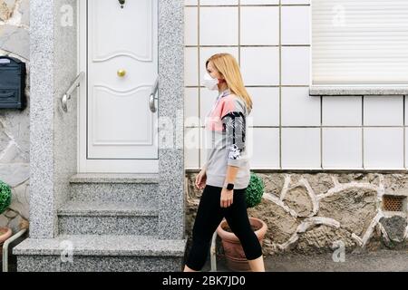 Una donna con una maschera sul suo volto cammina lungo la strada con una casa sullo sfondo Foto Stock