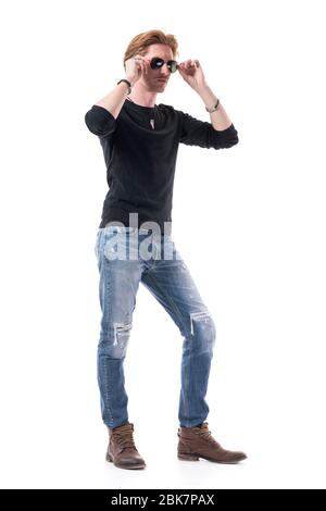 Fresco giovane bell'uomo con capelli rossi che indossano jeans strappati e camicia nera che prova su occhiali da sole. Corpo intero isolato su sfondo bianco. Foto Stock
