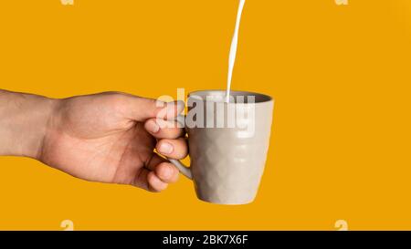 Giovane uomo irriconoscibile che tiene una tazza di bevanda calda con latte che vi versa, fondo arancione. Panorama Foto Stock