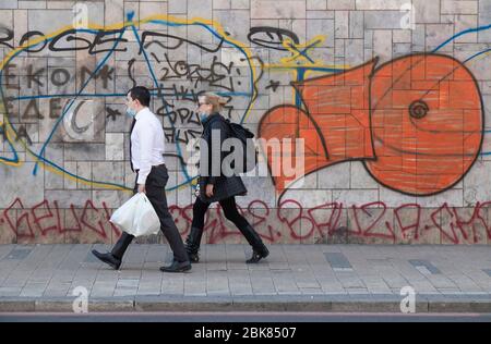 Belgrado, Serbia - 9 aprile 2020: Uomo e donna camminano lungo la strada della città indossando maschere chirurgiche e portando borse di alimentari Foto Stock