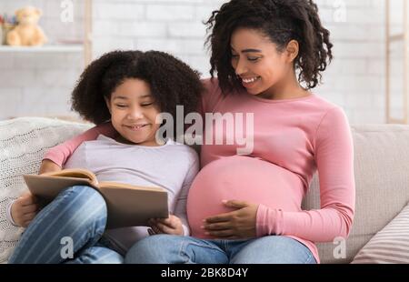 Carino ragazza piccola nera lettura Libro con mamma incinta a casa Foto Stock