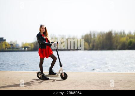 Giovane donna in giacca e abito in pelle tiene scooter elettrico Foto Stock