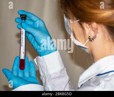 medico/scienziato femminile con maschera, tenendo e guardando il campione di sangue con guanti monouso blu, da dietro la scena Foto Stock