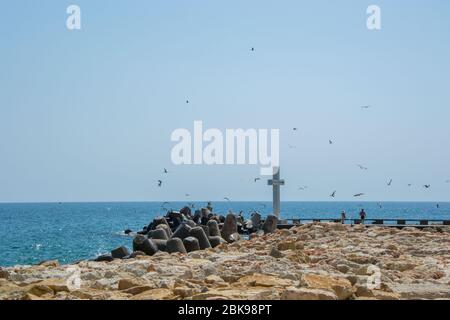 Mare, vista di una grande croce di cemento sulla baia, persone a piedi e uccelli volare, acque blu e cielo, frangiflutti, stagione estiva, natura Foto Stock