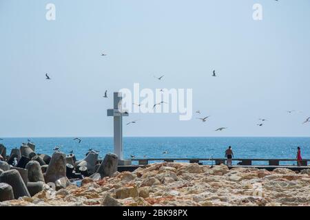 Mare, vista di una grande croce di cemento sulla baia, persone a piedi e uccelli volare, acque blu e cielo, frangiflutti, stagione estiva, natura Foto Stock