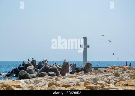 Mare, vista di una grande croce di cemento sulla baia, persone a piedi e uccelli volare, acque blu e cielo, frangiflutti, stagione estiva, natura Foto Stock