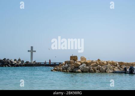 Mare, vista di una grande croce di cemento sulla baia, persone a piedi e uccelli volare, acque blu e cielo, frangiflutti, stagione estiva, natura Foto Stock