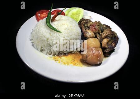 Tazza di riso bollito con brodo di pollo su un piatto bianco isolato su fondo nero. Riso cotto su un piatto bianco, peperoncino, pomodoro, cetriolo. Foto Stock