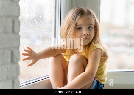 una bambina in una blusa gialla si siede accanto alla finestra con le gocce di pioggia ed è triste. i bambini sono annoiato a casa a causa della quarantena e di auto-isolamento Foto Stock