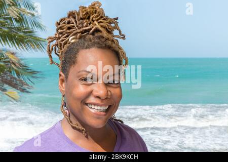 Close Up Ritratto di bella donna nera, africana, o carribea ridendo all'aperto che si affaccia sulla splendida spiaggia e l'oceano. Foto Stock