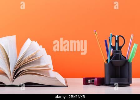 Un libro e un piedistallo con penne, matite e forbici su un tavolo di legno chiaro. Il libro è aperto, le foglie della pagina sono fanning out, un marrone arancio dorso Foto Stock