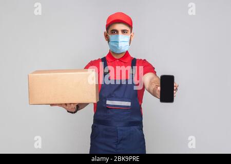 Consegna in quarantena. Questo è per voi! Giovane uomo con maschera chirurgica medica in uniforme blu e t-shirt rossa in piedi, tenendo scatola di cartone e spettacolo Foto Stock