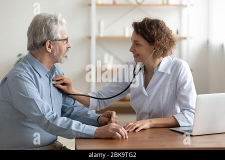 Medico femminile che ascolta il cuore del paziente maschio anziano usando lo stetoscopio Foto Stock