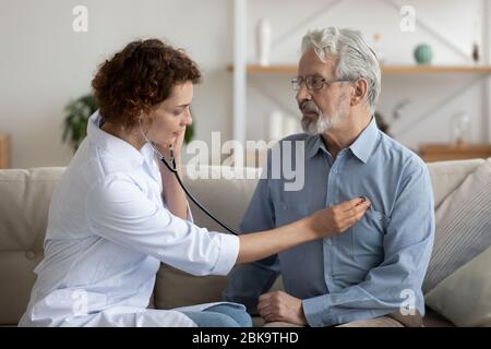 Medico femminile che tiene stetoscopio in ascolto paziente anziano durante la visita di homecare Foto Stock