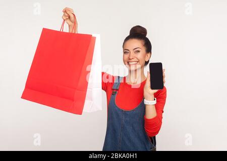 Ritratto di happy shopper, ragazza in tute denim tenendo borse e cellulare, pubblicità applicazione per l'ordine online, l'acquisto Foto Stock